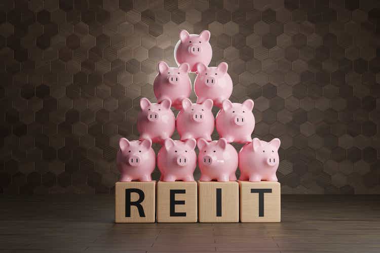 Куча розовых копилок на деревянных блоках, написанных алфавитом REIT.  Иллюстрация концепции инвестиционного траста недвижимости