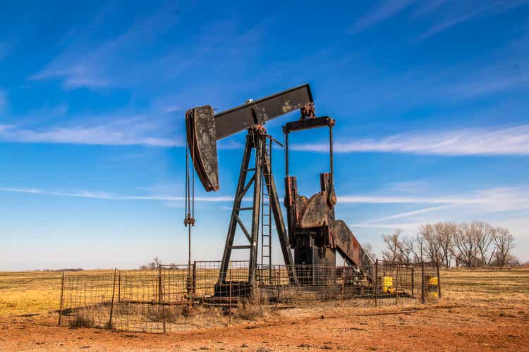 Большой старый ржавый насос для нефтяной скважины, окруженный панельным забором для скота в поле с очень голубым небом зимой.