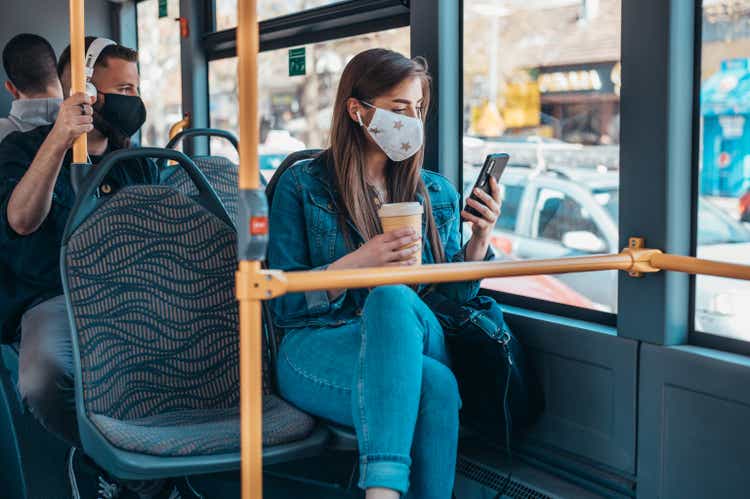 Женщина в защитной маске едет в автобусе и пользуется смартфоном
